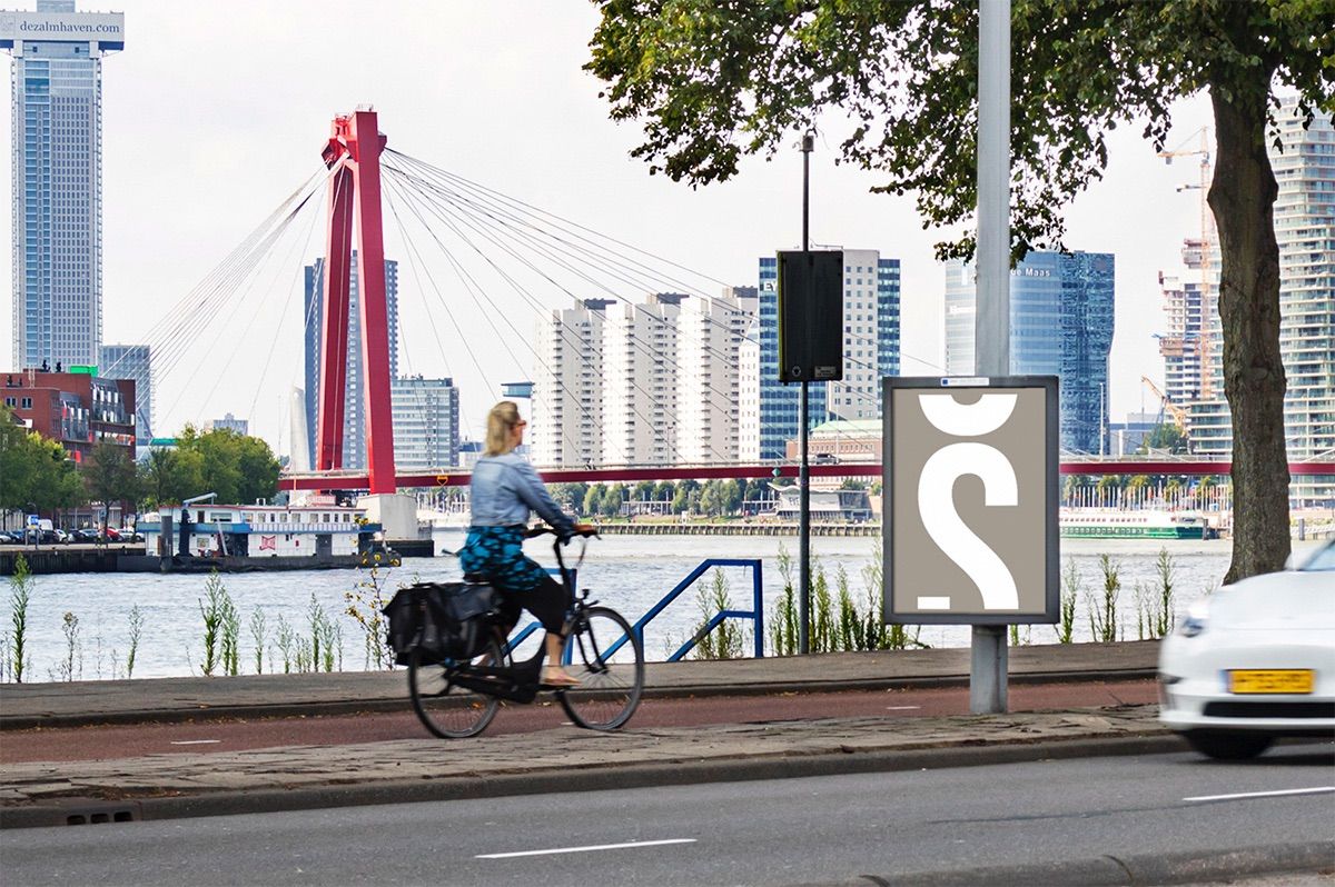 A0-poster aan een lantaarnpaal langs de Maas in Rotterdam, met een fietser en de Willemsbrug op de achtergrond