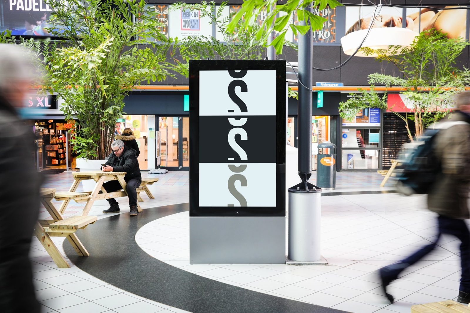 Digitaal scherm in abriformaat op een overdekt winkelcentrumplein met voorbijgangers